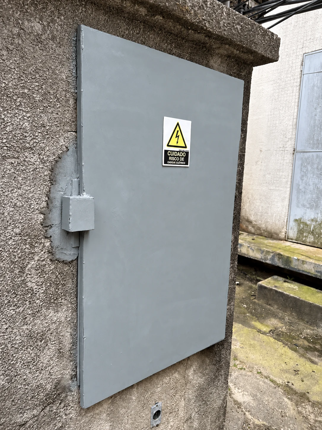 Porta metálica instalada, com proteção do fechamento e sinalização de risco elétrico.
